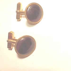 May include: A pair of gold-toned cufflinks with a dark purple, oval-shaped center stone. The cufflinks have a classic design, suitable for formal wear. The stones are set in a gold-colored bezel.