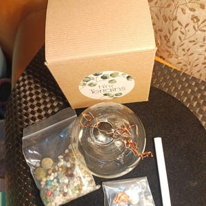 May include: A kit for creating a terrarium, featuring a clear glass globe, a bag of colorful stones, a small seashell, a brown cardboard box with the text "Tiny Terrains", and a white stick.