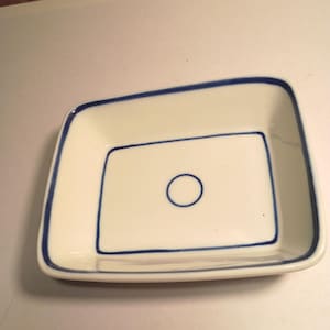 May include: A rectangular white ceramic dish with rounded corners, featuring a thin blue painted border along the rim and a matching blue rectangular frame with a small blue circle centered on the interior base of the shallow serving tray.