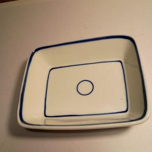 May include: A rectangular, off-white ceramic dish with a blue border and a blue inner rectangle. A blue circle is in the center of the inner rectangle. The dish has rounded corners and is likely for serving or display.