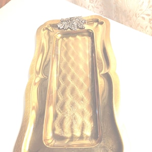 May include: A rectangular gold-toned serving tray with a decorative silver-colored grape cluster and leaf design. The tray has a slightly scalloped edge and a textured interior. Ideal for serving appetizers or displaying small items.