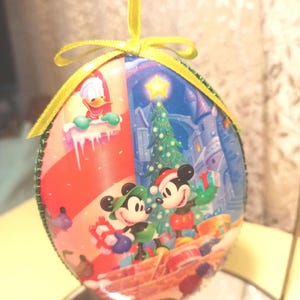 May include: A round Christmas ornament with a festive scene featuring Mickey Mouse and Minnie Mouse in front of a Christmas tree.  Donald Duck is featured in the top left corner of the ornament.  The ornament is decorated with a green ribbon.