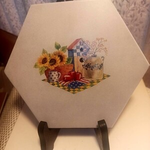 May include: A hexagonal decorative tile featuring a country-themed illustration. The artwork includes sunflowers, a birdhouse, a basket, apples, and a jug, set against a backdrop of an American flag-inspired design. The tile is displayed on a wooden stand.