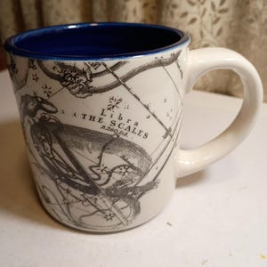 Peut inclure: Tasse en c&eacute;ramique blanche avec un int&eacute;rieur bleu et une anse incurv&eacute;e. La tasse pr&eacute;sente une illustration en noir et blanc de la constellation de la Balance et le texte "Libra The Scales". Une tasse d&eacute;corative pour les amateurs d'astrologie.