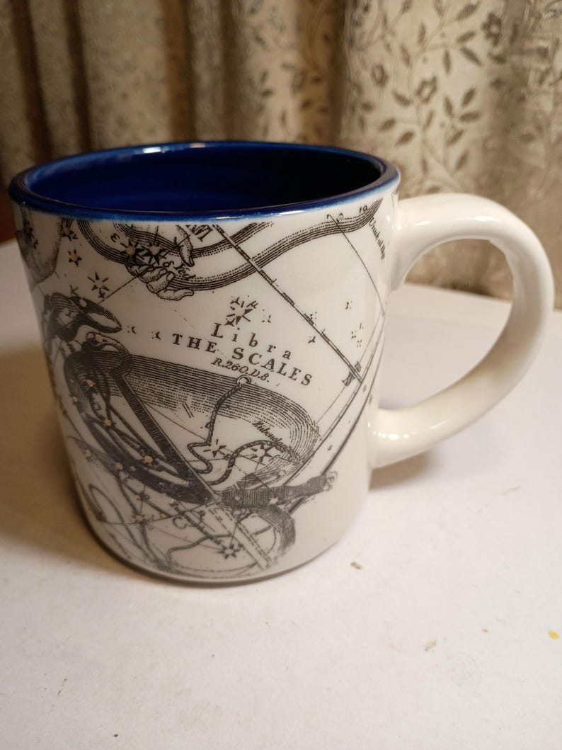 Peut inclure: Tasse en c&eacute;ramique blanche avec un int&eacute;rieur bleu et une anse incurv&eacute;e. La tasse pr&eacute;sente une illustration en noir et blanc de la constellation de la Balance et le texte "Libra The Scales". Une tasse d&eacute;corative pour les amateurs d'astrologie.