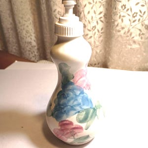 May include: A white ceramic soap dispenser with a floral design in shades of blue, pink, and green. The dispenser has a white plastic pump and a ribbed collar. The bottle is approximately 7 inches tall.