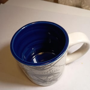 Peut inclure: Une tasse en c&eacute;ramique avec une anse blanche et un int&eacute;rieur bleu. L'ext&eacute;rieur pr&eacute;sente un motif de carte en tons gris. La tasse a une forme l&eacute;g&egrave;rement inclin&eacute;e et une finition lisse et brillante.