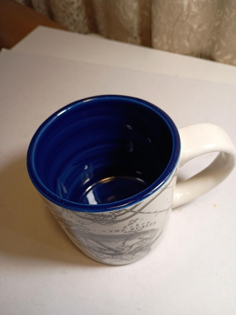 Peut inclure: Une tasse en c&eacute;ramique avec une anse blanche et un int&eacute;rieur bleu. L'ext&eacute;rieur pr&eacute;sente un motif de carte en tons gris. La tasse a une forme l&eacute;g&egrave;rement inclin&eacute;e et une finition lisse et brillante.