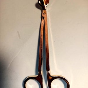 May include: A gold-colored candle wick trimmer with a circular blade and finger loops. The trimmer has long, slender handles and is designed to cut candle wicks cleanly. The tool is made of metal and has a shiny finish.
