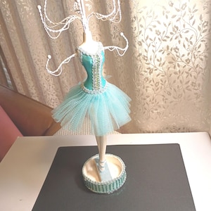 May include: A jewelry display stand shaped like a ballerina wearing a turquoise bodice with silver trim and a matching tulle tutu. The figure stands on a circular base with a light blue platform, featuring metal branches extending upward for hanging items.