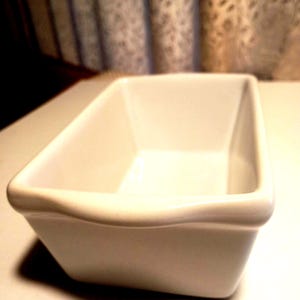 May include: A rectangular, off-white ceramic baking dish. The dish has a slightly curved rim and is empty. The background includes a patterned curtain.