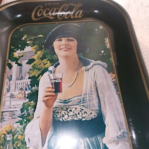 May include: Vintage Coca-Cola serving tray with a black border and a classic advertisement. The image features a woman in a white dress and hat holding a glass of Coca-Cola. The Coca-Cola logo is displayed prominently.