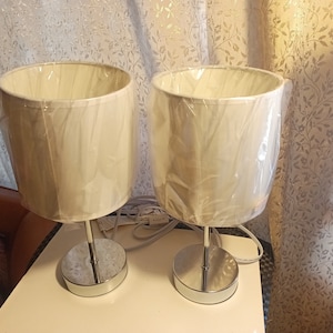 May include: Two white table lamps with chrome bases and white fabric shades. The lamps are on a white surface.