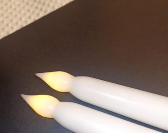 May include: Two white, battery-operated taper candles with a soft, flickering yellow light at the tips. The candles are lying on a dark surface, suggesting a decorative or ambient lighting theme.