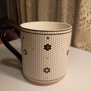 May include: A white ceramic mug with a black handle and rim. The mug features a honeycomb pattern with black and gold floral accents. The mug is designed for hot beverages.