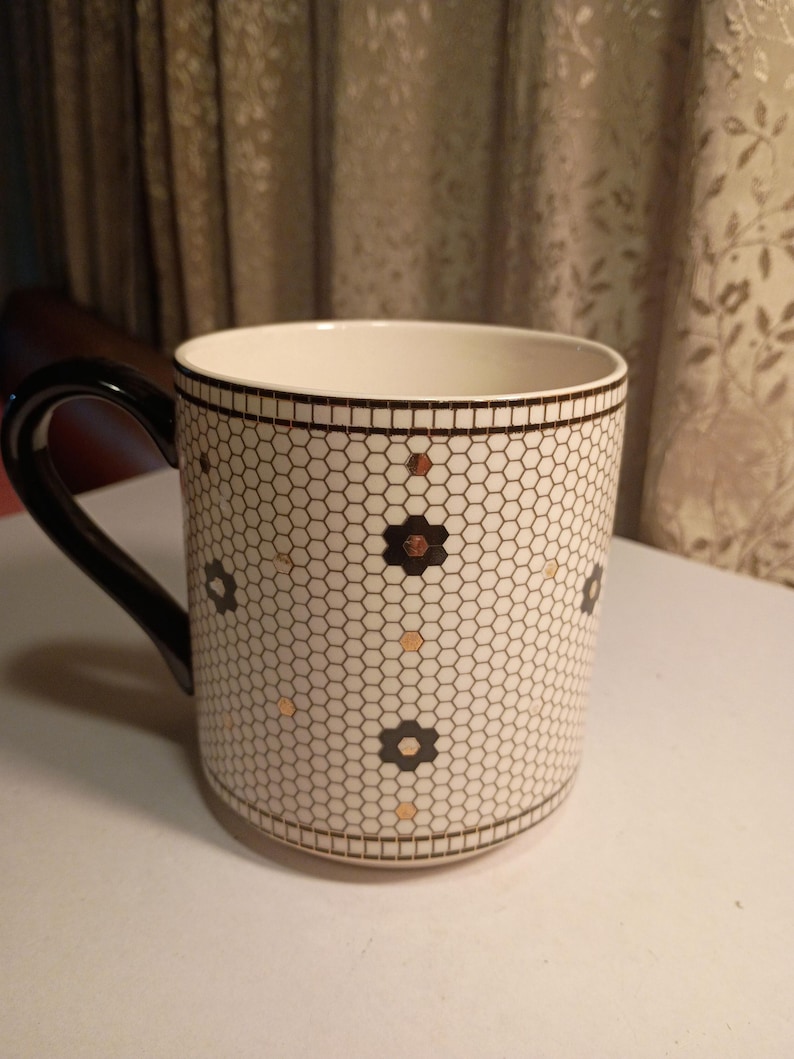 May include: A white ceramic mug with a black handle and rim. The mug features a honeycomb pattern with black and gold floral accents. The mug is designed for hot beverages.