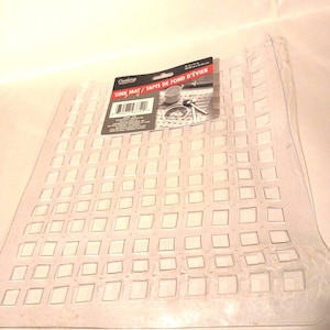 May include: A clear, rectangular plastic sink mat featuring a grid of square cutouts. A black product label is attached to the top, displaying the text "Cooking" and "SINK MAT / TAPIS DE FOND D'&Eacute;VIER" along with a barcode and dimensions of 12 in x 11 in.