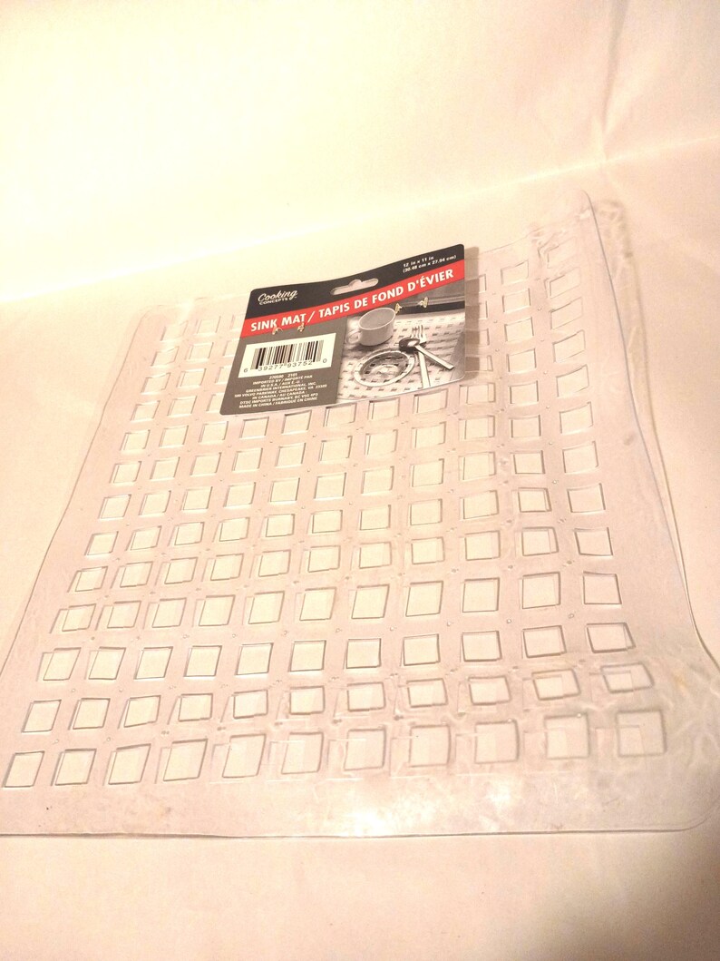 May include: A clear, rectangular plastic sink mat featuring a grid of square cutouts. A black product label is attached to the top, displaying the text "Cooking" and "SINK MAT / TAPIS DE FOND D'&Eacute;VIER" along with a barcode and dimensions of 12 in x 11 in.