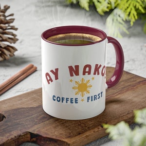 May include: White ceramic coffee mug with a red rim and handle. The mug has a graphic of a sun with a yellow center and red rays, a blue and red star on each side of the sun, and the text "Ay Nako Coffee First" in red and blue letters.