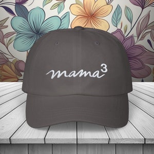 May include: A gray baseball cap with the word "mama³" embroidered in white script across the front. The hat is set against a floral patterned background.