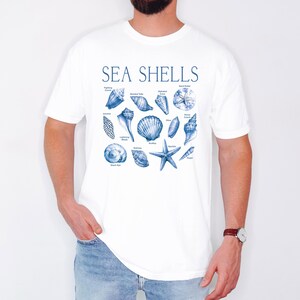 Op de afbeelding: Wit T-shirt met een blauwe illustratie van verschillende schelpen en de tekst "SEA SHELLS".