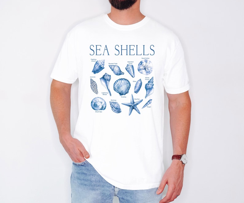 Op de afbeelding: Wit T-shirt met een blauwe illustratie van verschillende schelpen en de tekst "SEA SHELLS".