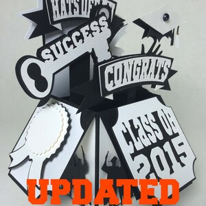 Op de afbeelding: Zwart-witte afstudeerkaart met een pop-up ontwerp. De kaart heeft een sleutel, een afstudeerpet, een banner met de tekst "Gefeliciteerd" en een banner met de tekst "Klas van 2015". De kaart heeft ook een lint en een cirkel met een lege ruimte voor een bericht. De tekst "GEUPDATEERD GRAD JAAR TOT 2026" is in oranje aan de onderkant van de kaart gedrukt.