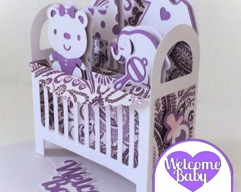 Puede incluir: Una tarjeta 3D pop-up de color morado y blanco con una cuna, una jirafa, un oso de peluche y un elefante. La tarjeta dice "Welcome Baby" en la parte delantera y "Welcome Baby" en la parte trasera.