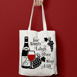 May include: White tote bag with black text and a graphic of a wine bottle, a glass of wine, and grapes. The text reads "Live Simply Laugh Often Wine A Lot".