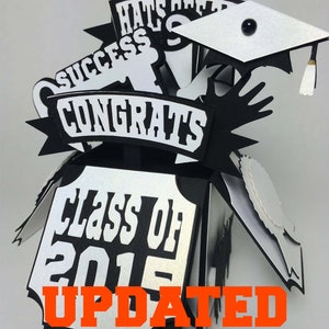 Op de afbeelding: Zwarte en witte pop-up box met een afstudeerthema met een afstudeerpet, een sleutel en een banner met de tekst "Gefeliciteerd". De doos is versierd met de woorden "Hats Off" en "Success". De doos heeft ook een zilveren plaquette met de tekst "Klas van 2015" met de tekst "UPDATED GRAD YEARS TO 2026" in oranje eronder.
