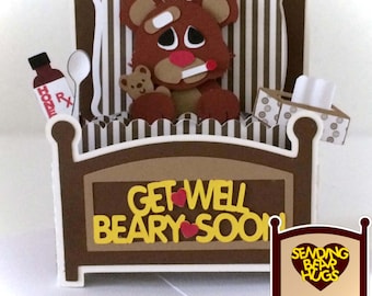 May include: A brown teddy bear wearing a bandage and a thermometer is lying in bed with a tissue box and a bottle of honey. The bed is brown with white and brown striped sheets. The card reads "Get Well Beary Soon" and the back of the card reads "Sending Bear Hugs".