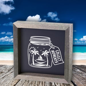 Può includere: Un cartello in ardesia incorniciato con una silhouette bianca di un barattolo di vetro con palme e una barca a vela all'interno. Il barattolo ha un'etichetta che pende da esso che dice "Collect Moments".