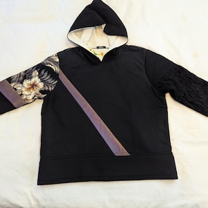May include: Black hoodie with a contrasting floral print on one sleeve and a textured sleeve on the other. The hoodie features a hood with a cream-colored lining and a diagonal stripe across the front.