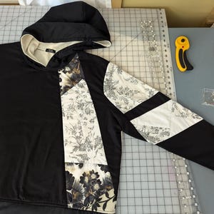 May include: A black hooded sweatshirt with a patchwork design featuring floral and geometric patterns. The sweatshirt has a white hood lining and a white zipper pull.