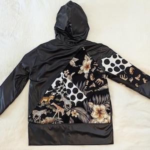 May include: Black hooded jacket with a patchwork design featuring black and white polka dots, tropical flowers, and animal prints. The jacket has a diagonal seam and a black lining.