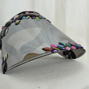 May include: A black face shield with a silver mirrored visor and a decorative border of colorful rhinestones.