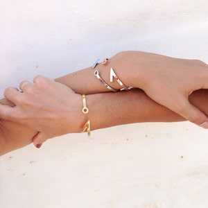 May include: Three gold bracelets on a person's wrist. The bracelets are all different styles, one is a simple band, one is a curved shape, and the other is a more intricate design with a circle in the center.