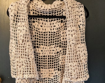 May include: A beige sleeveless crochet vest featuring an openwork grid pattern with integrated floral motifs. The garment has a draped, open front design with a scalloped edge and is constructed from a light-colored yarn in a repeating stitch.