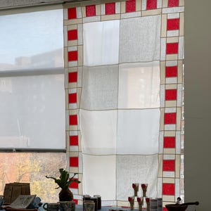 May include: A window curtain with a geometric design of white, gray, and red squares. A table is set with various items, including cups, jars, wooden spoons, and a small plant, all arranged on a blue tablecloth.