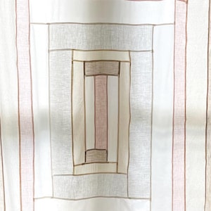 May include: Cream-colored fabric with a geometric design. The pattern features vertical stripes in shades of beige, pink, and brown, with a central rectangular design. The fabric appears to be lightweight and suitable for curtains or other home decor items.