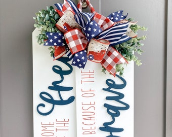 Könnte beinhalten: Ein weißes Holzschild mit dem Text "Home of the Brave because of the Free" in roten und blauen Buchstaben. Das Schild ist mit einer rot-weiß-blauen Schleife und Grünpflanzen verziert.