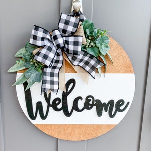 May include: A round wooden door hanger with a white and brown striped design. The word "Welcome" is written in black script on the white stripe. A black and white checkered bow with burlap ribbon and green leaves is attached to the top.