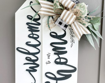 Op de afbeelding: Een wit houten bord met zwarte letters met de tekst "Welcome to our home" met een zwart-wit geblokt lint, een bruin gestreept lint en groene bladeren.