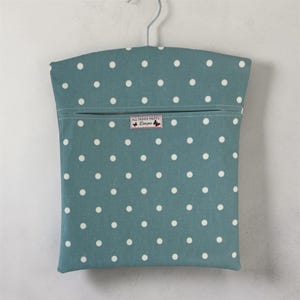 Peut inclure: Un sac à vêtements à pois turquoise avec une fermeture éclair et un cintre en métal. Le sac présente des pois blancs et une étiquette indiquant "All Things Pretty Designs".