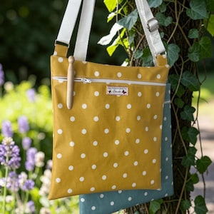 Pode incluir: Uma bolsa tiracolo amarelo-mostarda com bolinhas brancas, apresentando uma alça ajustável branca, um bolso frontal com zíper e uma pequena etiqueta retangular com a inscrição All Things Pretty Designs. Um pino de madeira clara está preso ao zíper.