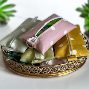 May include: A collection of small rectangular zipped pouches in dusty pink, mustard yellow, and sage green, each featuring a white polka dot pattern. Each pouch includes a white zip closure and a gold-toned metal D-ring attached to one side.