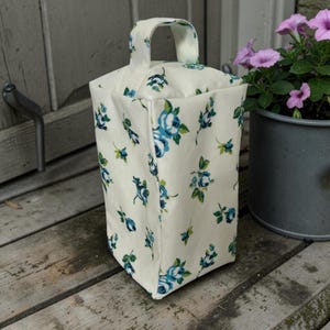 May include: A cream-coloured fabric doorstop featuring a repeating pattern of blue roses and green leaves. The doorstop has a rectangular shape with a sturdy fabric handle attached to the top centre, allowing for easy movement and placement on the floor.