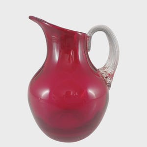 Peut inclure: Une carafe en verre rouge rubis avec une poignée claire et incurvée. La carafe a un corps arrondi et un bec verseur. La poignée est fixée au sommet de la carafe et se courbe vers le bas pour rejoindre le corps.