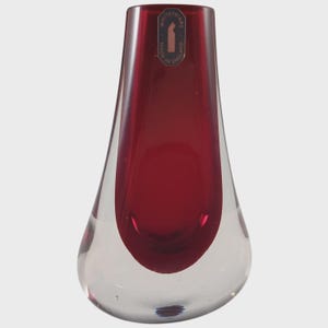 May include: A teardrop-shaped glass vase with a clear base and a vibrant red upper section. The vase has a smooth, polished surface and a label that reads "Made in England". The design is simple and elegant.
