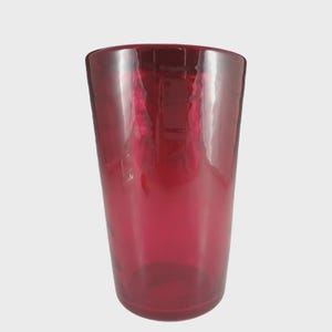 May include: A tall, cylindrical, ruby-red glass. The glass has a smooth, glossy surface and a slightly flared rim. The transparent material allows light to pass through, creating highlights and reflections. The glass is empty and stands against a neutral background.